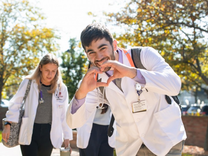 A student in lab coat holds his hands together in a heart shape on his way to class 