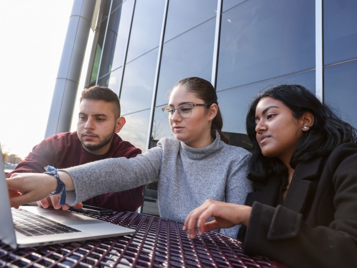 Three students gather around laptop at a picnic table outside and discuss coursework