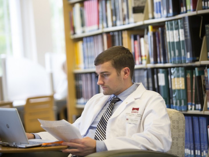 Student in white coat sitting in chair in front of the bookshelf doing work on their laptop