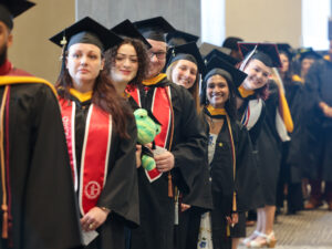 Graduates stand in line with commencement regalia smiling
