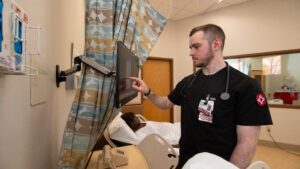 Robert Bautz, a BSN student, presses the controls of an IV monitor as he works bedside with a patient