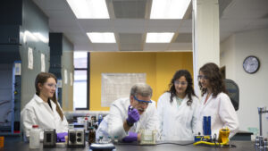 Students and faculty dressed in lab safety gear examine samples while using a pipette