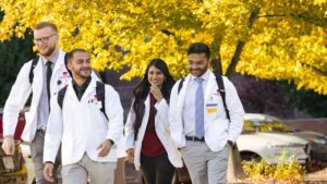 A group of pharmacy students in white coats walk together on a sunny fall day