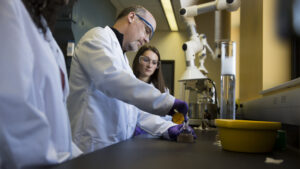 Students observing professor measuring chemical reagents with a graduated cylinder in lab safety gear